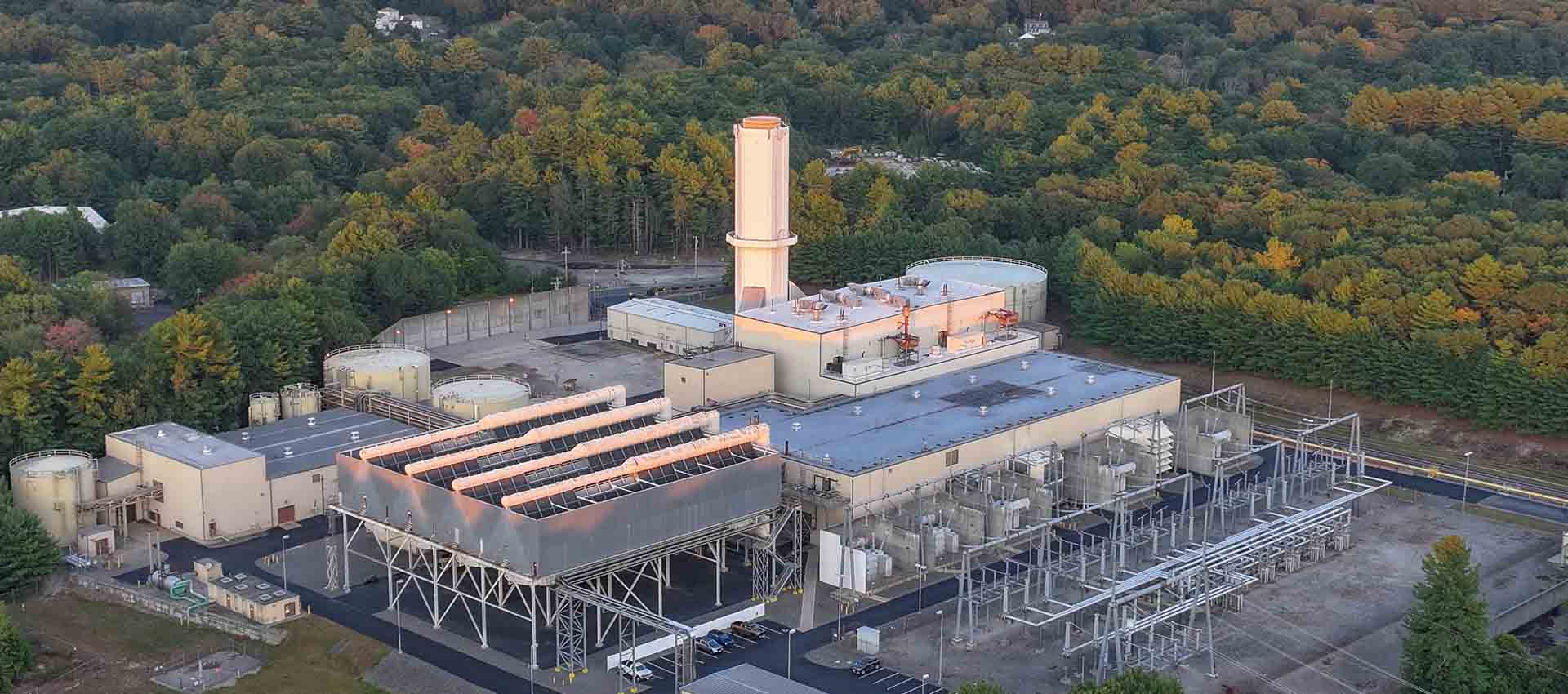 Aerial view of a power plant surrounded by dense forest under a soft sunset sky, with industrial structures and a tall central stack visible.