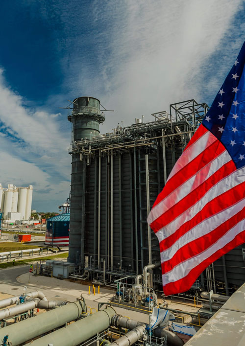 American flag at a NextEra Energy facility with storage tanks and piping under blue sky.