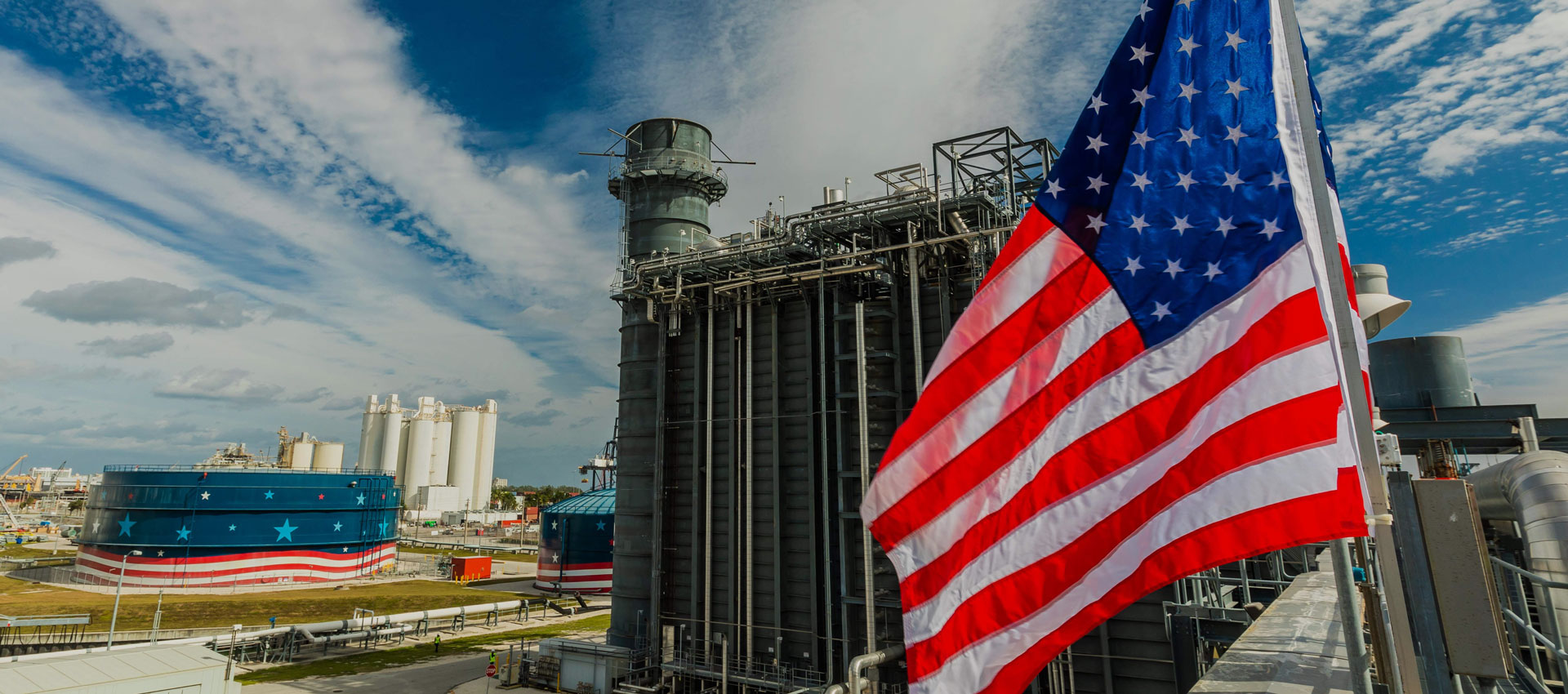 American flag at a NextEra Energy facility with storage tanks and piping under blue sky.