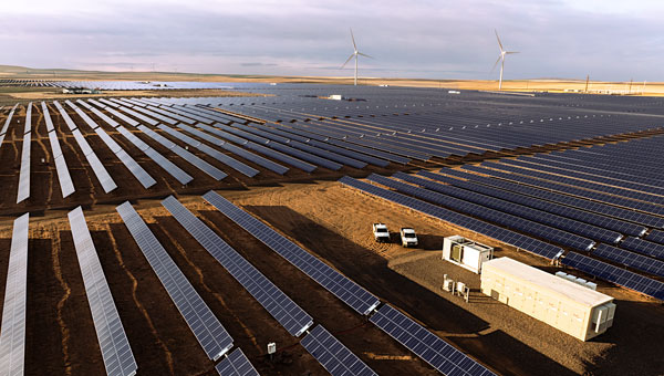 A large solar farm with rows of solar panels, abattery energy storage unit and wind turbines in the background under a cloudy sky.