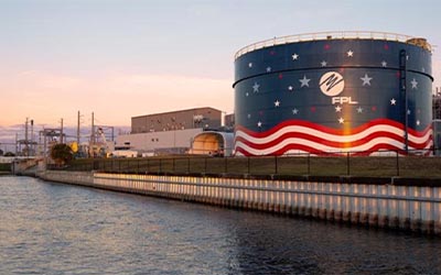 A waterfront energy facility featuring a large fuel storage tank wrapped in an American flag mural and FPL logo, with power plant infrastructure visible in the background at sunset