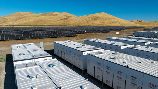 Battery storage units and expansive solar panel array near grassy hills.